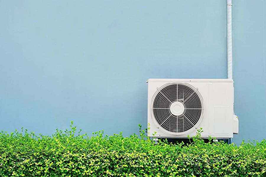 A heat pump against a blue wall above a green hedge